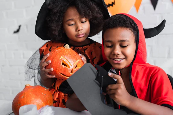 Happy african american boy in halloween costume cutting carton with scissors near sister with carved pumpkin — Stock Photo
