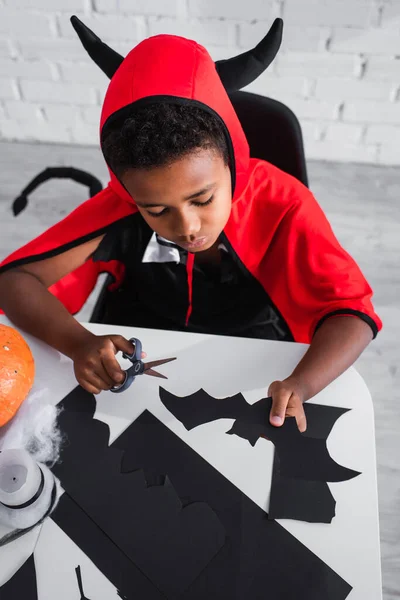 High angle view of african american boy in halloween costume holding scissors and paper cut bat — Stock Photo