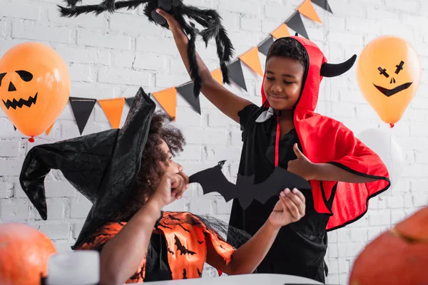 Africano americano ragazza in halloween costume tenendo carta tagliata pipistrello vicino felice fratello con giocattolo ragno mostrando pollice su — Foto stock