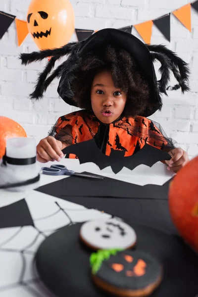 Spooky african american girl in pointed witch hat holding paper cut bat — Stock Photo