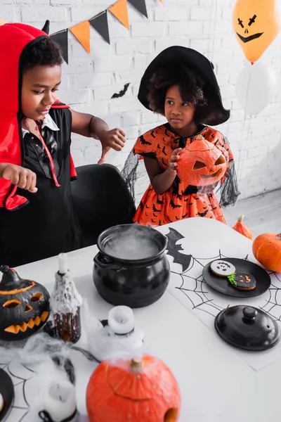 Menino americano africano em traje de Halloween mostrando polegares para baixo enquanto prepara poção perto da irmã — Fotografia de Stock