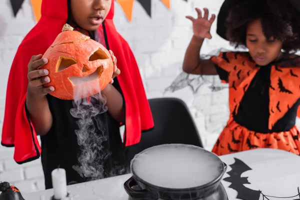 Africano americano chico en halloween traje celebración tallada calabaza con humo mientras preparación poción cerca borrosa hermana - foto de stock