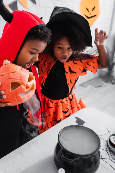 Menino americano africano em traje de Halloween segurando abóbora esculpida com fumaça enquanto prepara poção perto da irmã em chapéu pontiagudo — Fotografia de Stock