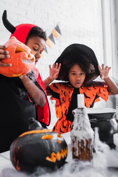 Africano americano ragazzo in halloween costume tenendo intagliato zucca mentre si prepara pozione vicino spettrale sorella in cappello a punta — Foto stock