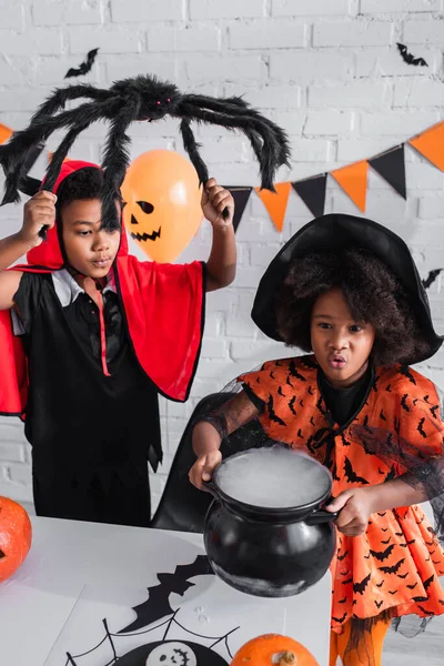 Criança americana africana em traje de Halloween segurando caldeirão bruxa com poção perto do irmão com aranha de brinquedo — Fotografia de Stock