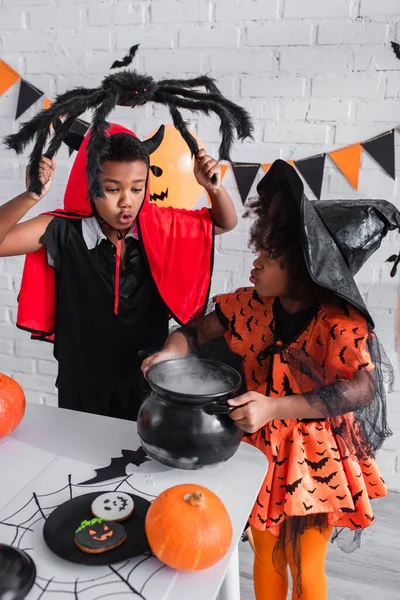 African american girl in pointed hat holding witch cauldron with potion near brother with toy spider — Stock Photo