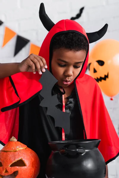 African american boy in devil halloween costume holding paper cut bat above cauldron — Stock Photo