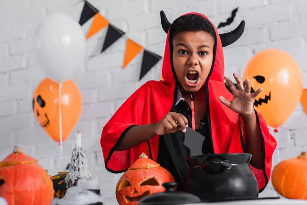 Spooky african american boy in devil halloween costume holding paper cut bat above cauldron — Stock Photo