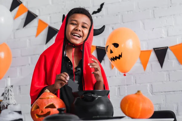 Happy african american boy in devil halloween costume holding paper cut bat above cauldron — Stock Photo
