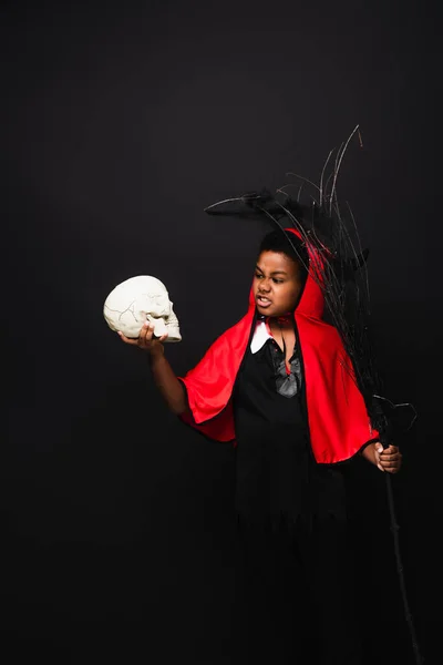 Scary african american boy holding broom and looking at skull isolated on black — Stock Photo