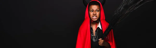 Irritated african american boy holding broom and grinning isolated on black, banner — Stock Photo