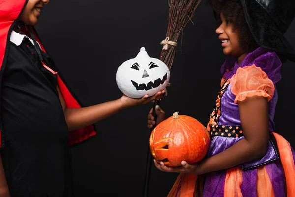 Niños afroamericanos sonrientes con disfraces de Halloween sosteniendo calabazas aisladas en negro - foto de stock