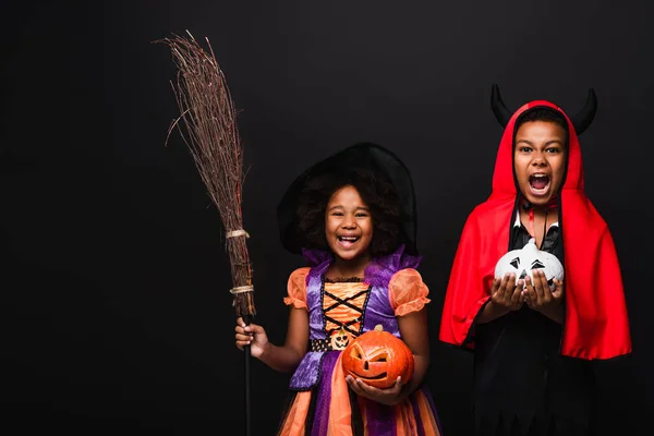 Animado afro-americano crianças em halloween trajes segurando esculpida abóboras isolado no preto — Fotografia de Stock