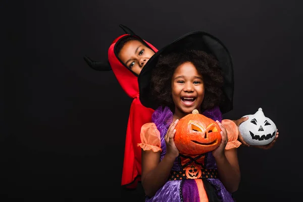 Alegre afroamericano niños en halloween trajes celebración espeluznante calabazas aislado en negro - foto de stock