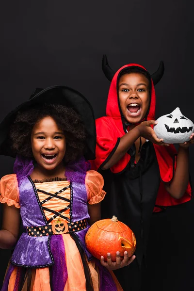 Cheerful african american children in halloween costumes holding spooky pumpkins isolated on black — Stock Photo