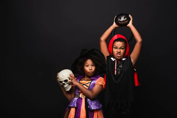 Assustador crianças afro-americanas em trajes halloween segurando crânio e abóbora esculpida isolado no preto — Fotografia de Stock