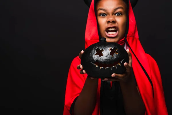 Spooky african american boy in devil costume holding dark carved pumpkin isolated on black — Stock Photo