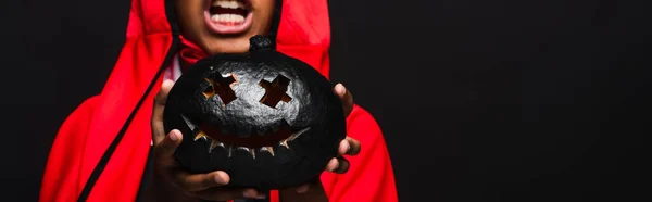 Recortado vista de espeluznante afroamericano chico en traje sosteniendo oscuro tallado calabaza aislado en negro, bandera - foto de stock