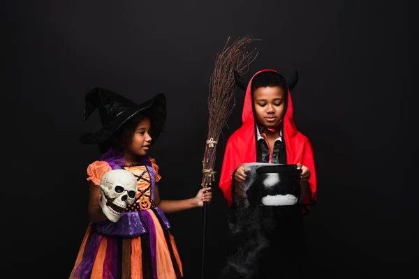 Афроамериканські діти в костюмах Хеллоуїна тримають череп і відьму котел зі зі зіллям ізольовані на чорному — стокове фото