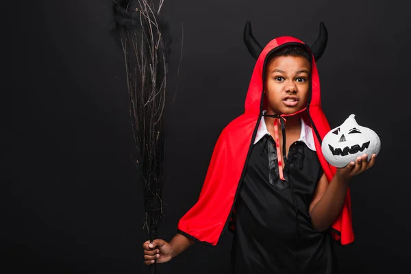 Spooky african american kid holding broom and drawn pumpkin isolated on black — Stock Photo