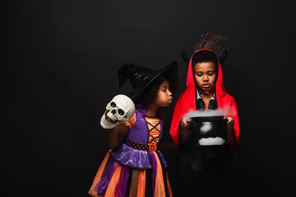 Africano americano chica en halloween traje celebración cráneo y soplando en bruja caldero con poción cerca hermano aislado en negro - foto de stock