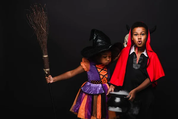 Afro-américaine fille dans halloween costume tenant balai et souffler sur chaudron avec potion près frère isolé sur noir — Photo de stock