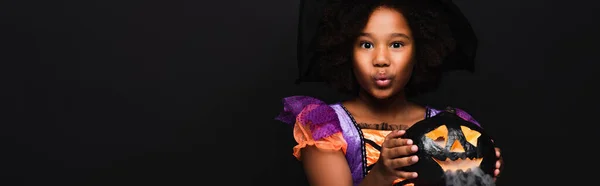Chica afroamericana en traje de halloween sosteniendo calabaza tallada aislada en negro, bandera - foto de stock
