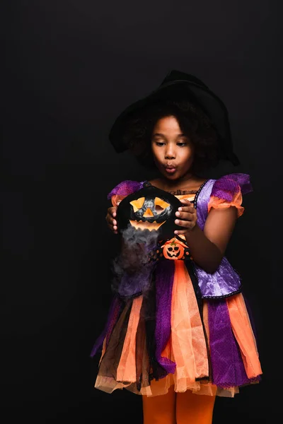 Chica afroamericana en traje de halloween mirando calabaza tallada con humo aislado en negro - foto de stock