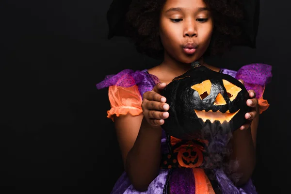 Surprised african american girl in halloween costume looking at carved pumpkin with smoke isolated on black — Stock Photo