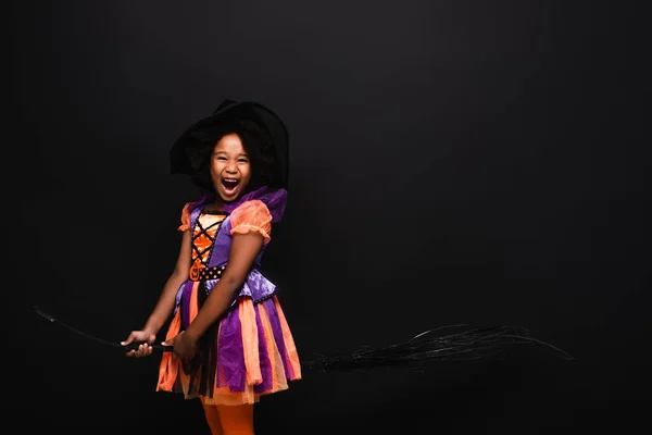 Excité afro-américaine fille en costume d'Halloween tenant balai isolé sur noir — Photo de stock