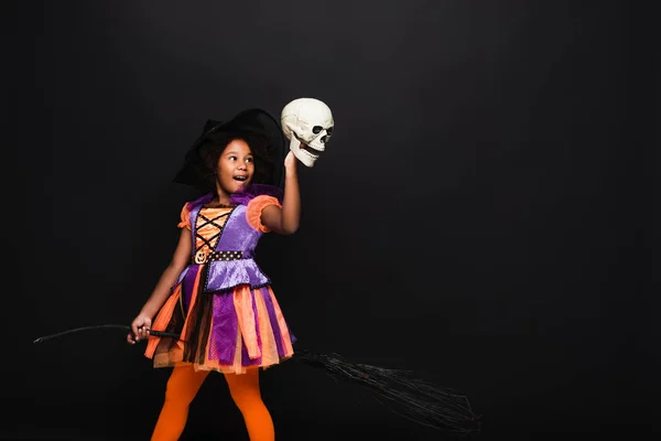 Excitado afroamericano chica en traje de bruja halloween en la parte superior de escoba sosteniendo cráneo aislado en negro - foto de stock