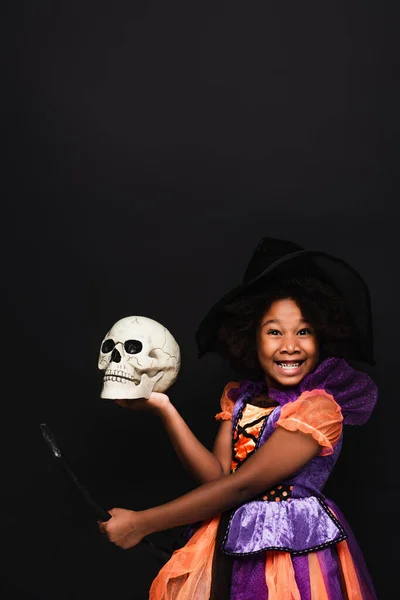 Cheerful african american girl in witch halloween costume on top of broom holding skull isolated on black — Stock Photo