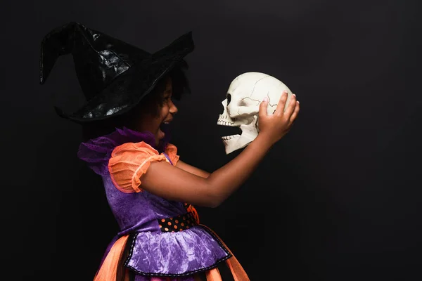Vista lateral da menina americana africana animado em bruxa halloween traje segurando crânio isolado no preto — Fotografia de Stock