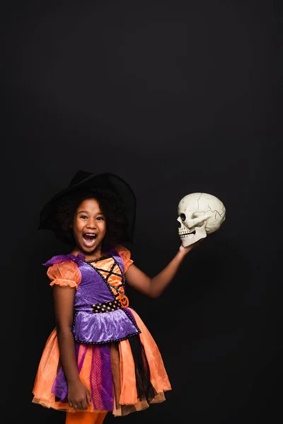 Asombrado afroamericano chica en bruja halloween traje celebración cráneo aislado en negro - foto de stock