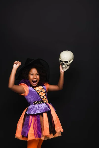 Здивована афроамериканська дитина в костюмі відьми Хелловін тримає череп ізольовано на чорному — стокове фото