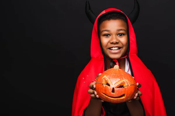Alegre afro-americano menino no diabo halloween traje segurando esculpida abóbora isolado no preto — Fotografia de Stock