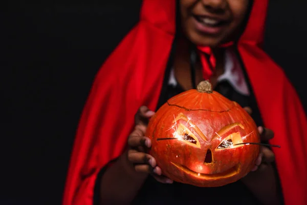 Cropped view of blurred and happy african american boy in devil halloween costume holding carved pumpkin isolated on black — Stock Photo