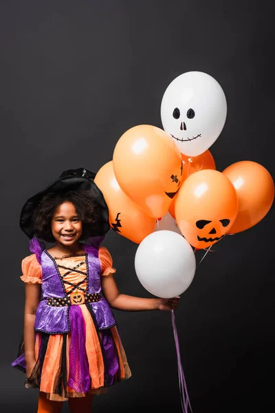 Glücklich afrikanisch-amerikanisches Mädchen im Halloween-Kostüm mit Luftballons auf schwarz isoliert — Stockfoto