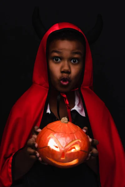 Assustador afro-americano menino no diabo halloween traje segurando esculpida abóbora isolado no preto — Fotografia de Stock