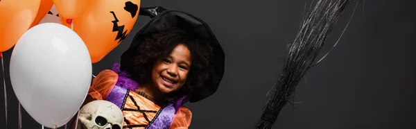 Happy african american girl in halloween costume holding broom and balloons isolated on black, banner — Stock Photo