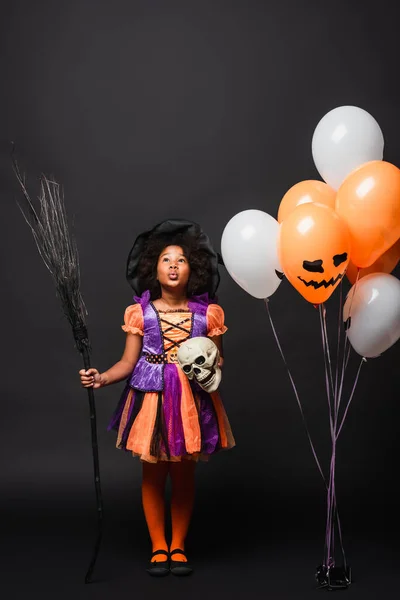 Surprised african american girl in halloween costume holding broom and skull near balloons isolated on black — Stock Photo