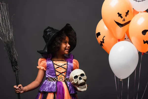 Menina americana africana alegre em traje halloween segurando vassoura e crânio perto de balões isolados em preto — Fotografia de Stock