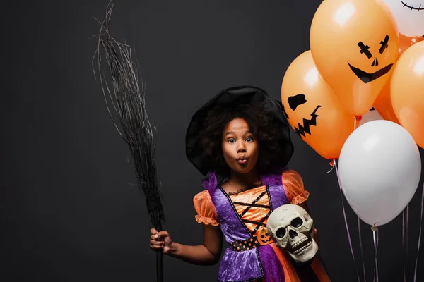 Menina americana africana em traje de bruxa halloween segurando vassoura e crânio perto de balões isolados em preto — Fotografia de Stock