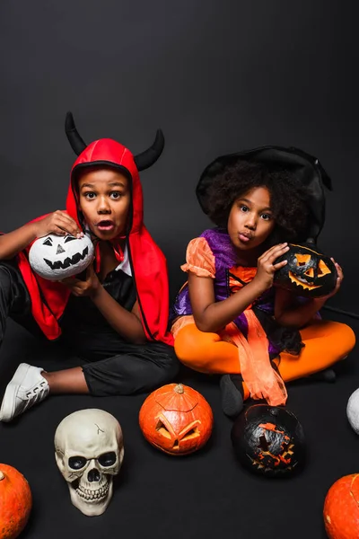 Assustador afro-americano crianças em halloween trajes segurando crânio e vassoura perto de abóboras em preto — Fotografia de Stock