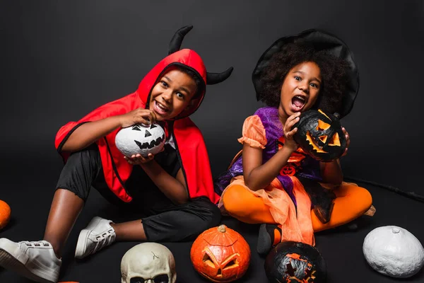 Irmãos africanos americanos em trajes de Halloween segurando abóboras esculpidas em preto — Fotografia de Stock