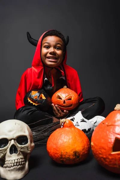 Menino americano africano feliz no traje do diabo halloween segurando abóboras esculpidas perto do crânio no preto — Fotografia de Stock