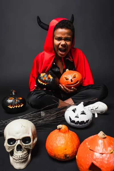 African american boy in halloween devil costume screaming while holding carved pumpkins near skull on black — Stock Photo