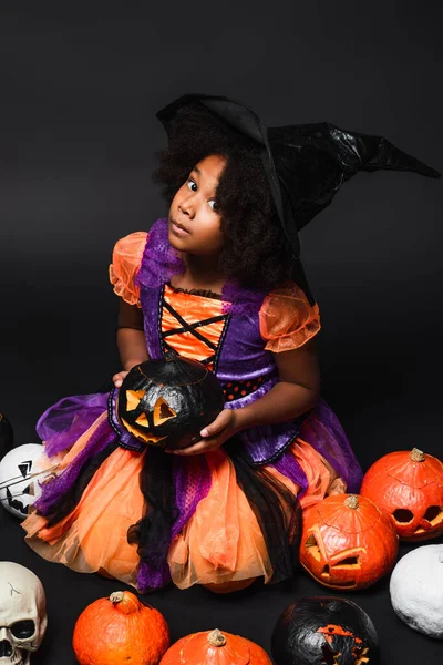 Vista de ángulo alto de chica afroamericana en traje de bruja y sombrero puntiagudo con calabaza tallada en negro - foto de stock