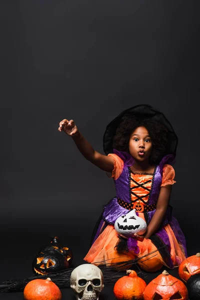Дивна афроамериканська дівчина в костюмі відьми і загострений капелюх біля різьблених гарбузів на чорному — стокове фото