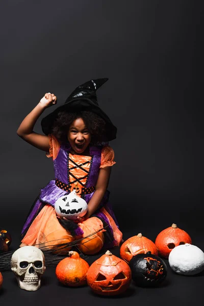 Excited african american child in witch costume and pointed hat near carved pumpkins on black — Stock Photo